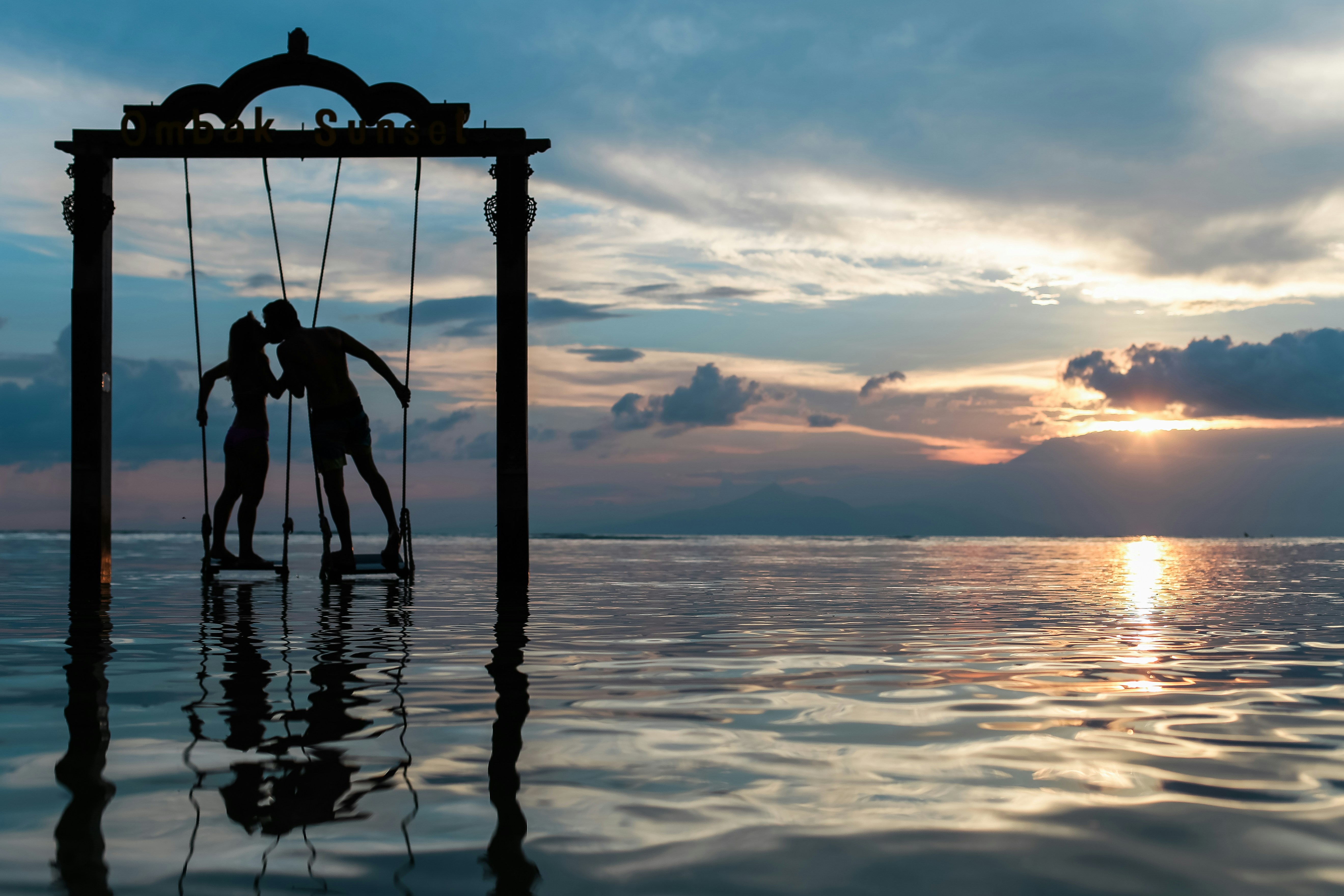 Image contains a couple standing on swings and kissing. Swings are in the middle of water at sunset with a cloudy dusk sky