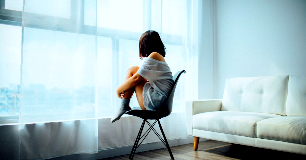 Living room with a white couch and a woman in white sitting on a chair in front of a large window with panels, looking out